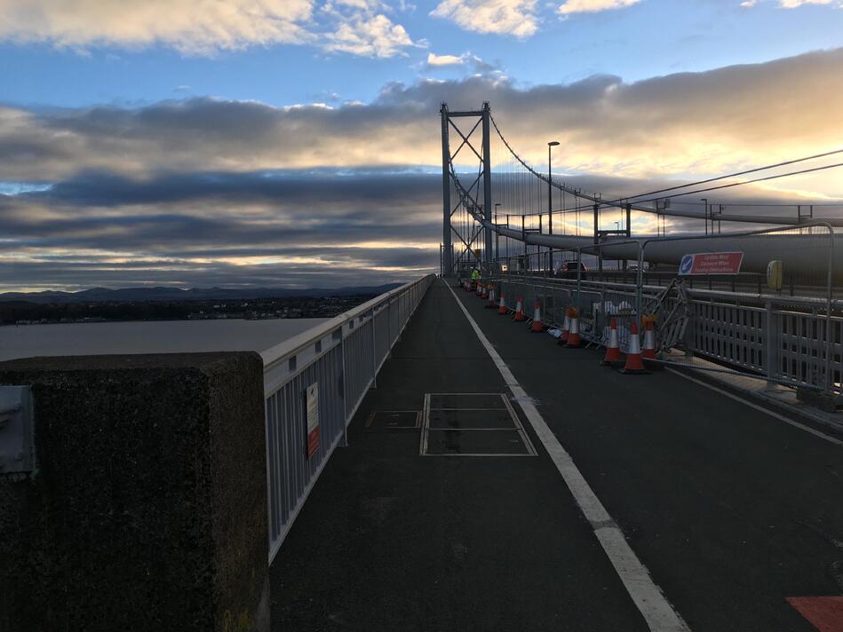 Looking onto a bridge, with a path directly ahead, fences and roadworks to the right, and water below on the left.