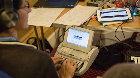 A person with their hands on a special “stenographic” keyboard, looking up to hear someone talking.